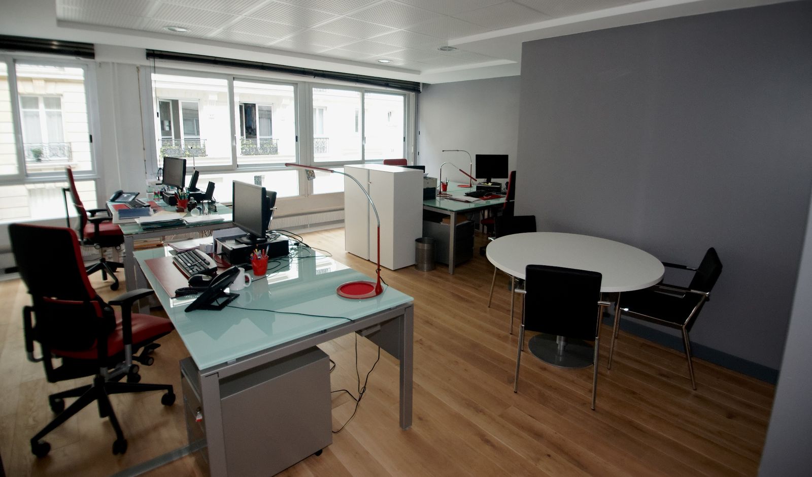 Plateau de bureaux : postes en verre, sièges rouges, table ronde de réunion, grandes fenêtres.